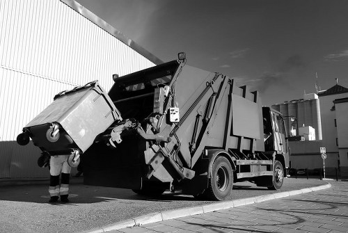 A large industrial rubbish collection truck is shown in a paved outdoor area, with its rear compaction mechanism actively lifting and emptying a bulk waste bin. The bin is made of metal with a corrugated surface and is positioned at an angle, partially lifted to empty its contents into the truck's hopper. The scene is set against a high, light-colored industrial building with vertical paneling, and the sky above is partly cloudy. Nearby, a person in work clothing with reflective elements is visible, guiding the bin into position. The environment appears to be a commercial or industrial waste management site, consistent with services provided by Commercial Waste Leatherhead, near the Leatherhead area, facilitating the disposal of construction or commercial waste as part of rubbish removal operations.