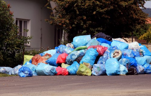 A large pile of discarded rubbish bags scattered along the edge of a residential street, with various shades of blue, red, yellow, green, and clear plastic, some filled with what appears to be household waste, on the pavement near a white residential building with a window and a garden area. The rubbish pile extends across the foreground, with a backdrop of trees and residential houses, indicating the area may be awaiting collection by a local waste removal service. The scene is illuminated by daylight, highlighting the crumpled textures and different materials of the bags, and reflecting the typical outdoor waste storage associated with rubbish collection in Leatherhead. Commercial Waste Leatherhead, a professional waste management company, is involved in handling such waste disposal needs within the local postcode region.