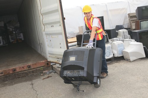 A person wearing a yellow safety helmet, a high-visibility vest with red and yellow reflective stripes, and gloves is standing outside a storage container, pushing a black electronic waste item towards the entrance. The electronic waste appears to be an old computer monitor or similar device, with a grey and black plastic casing and a curved screen. Behind the person, there are stacks of discarded monitors and other electronic appliances arranged on the concrete ground of a commercial or industrial area in Leatherhead, Surrey. The container’s open door reveals a dim interior with various stored materials, and part of the adjacent brick foundation is visible. The scene is well-lit with natural daylight, highlighting the textures of the electronic waste, the safety gear, and surrounding environment, demonstrating typical activity related to rubbish collection and disposal services, as offered by Commercial Waste Leatherhead, in the local postcodes around the town.