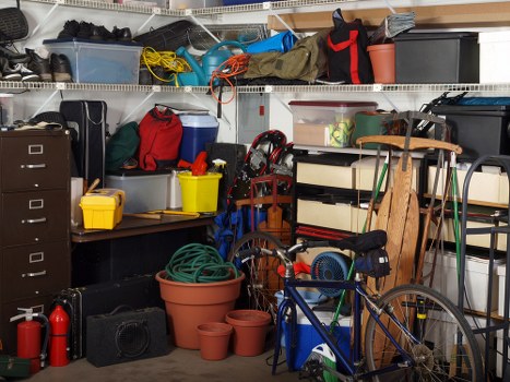 The image shows a cluttered storage area or garage filled with various household items and rubbish. In the foreground, there are several plastic plant pots of different sizes, some stacked, made of terracotta and plastic, with a garden hose coiled on top of a large terracotta pot. To the right, a blue bicycle with black pedals and a black saddle is leaning against a wooden shelving unit, which holds items such as a small black container, a roll of tape, and miscellaneous objects. Towards the back, a wooden chair with a curved backrest and slatted seat stands next to a white shelving unit containing various boxes, containers, and supplies, some of which are black or grey plastic storage boxes. On the left, a black filing cabinet with four drawers is visible, partially obscured by other items. Above, metal shelving runs across the width of the space, holding bags, tools, and miscellaneous equipment, including a red and black bag, a grey box, and cords. The overall scene depicts a typical cluttered garage or storage room ready for rubbish removal, with tools, garden supplies, and household clutter arranged haphazardly in a confined environment, indicative of a typical rubbish clearance in Leatherhead area.