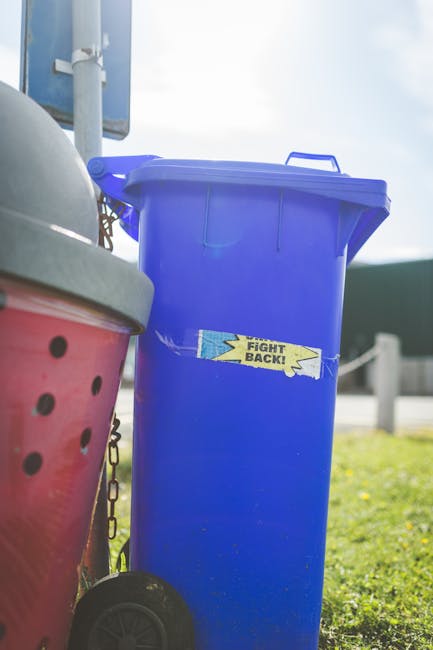 A row of large, rectangular wheelie bins lined up alongside a brick wall on a pavement, each with a plastic lid in different colors including green, red, blue, and black. The green bin in the foregrou