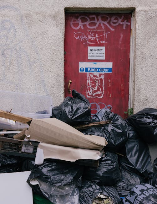 The image depicts the rear exterior wall of a brick building, featuring multiple air conditioning units and ventilation pipes mounted along the top section. Below, three large, wheeled rubbish bins ar