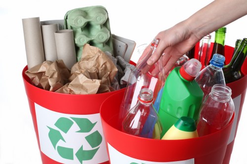 The image displays two red recycling bins with white labels featuring green recycling symbols; the left bin contains multiple white cardboard tubes, crumpled brown paper, and a green egg carton, while the right bin is filled with various clean plastic bottles in clear, green, and blue, with some having coloured caps, and is being handled by a person's hand. The setting appears to be indoors, with a clean, well-lit environment suitable for waste sorting. This visual emphasizes the separation of recyclable packaging and bottles, aligning with rubbish removal services offered by Commercial Waste Leatherhead in the area of Leatherhead, Surrey, post code KT22, supporting local waste management efforts.